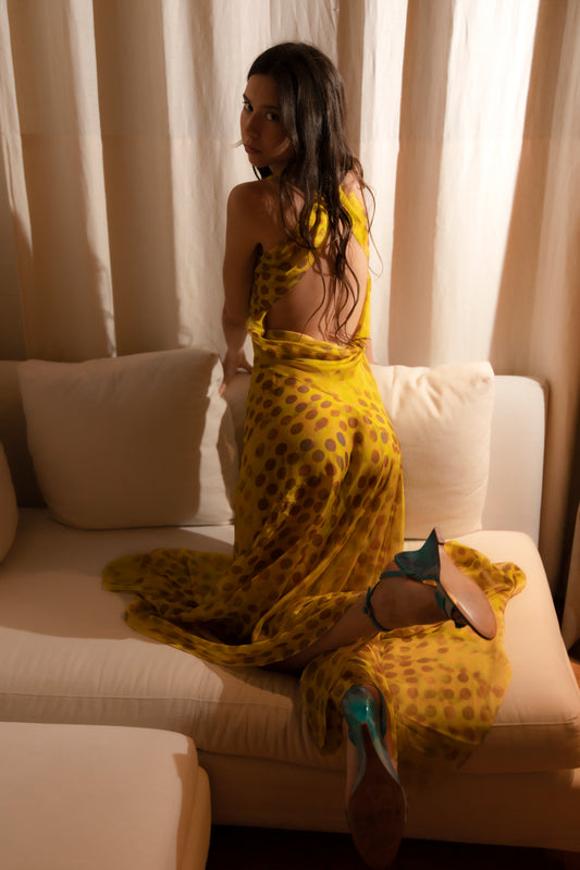 Woman in Romantica dress in Honeybaby sitting on a couch with curtains in the background