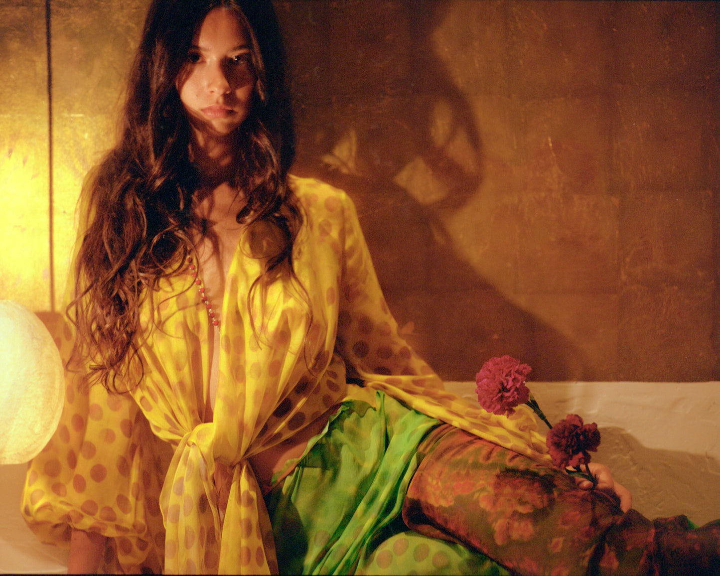 Woman in Electrica blouse in Honeybaby, sitting on a couch with flowers.