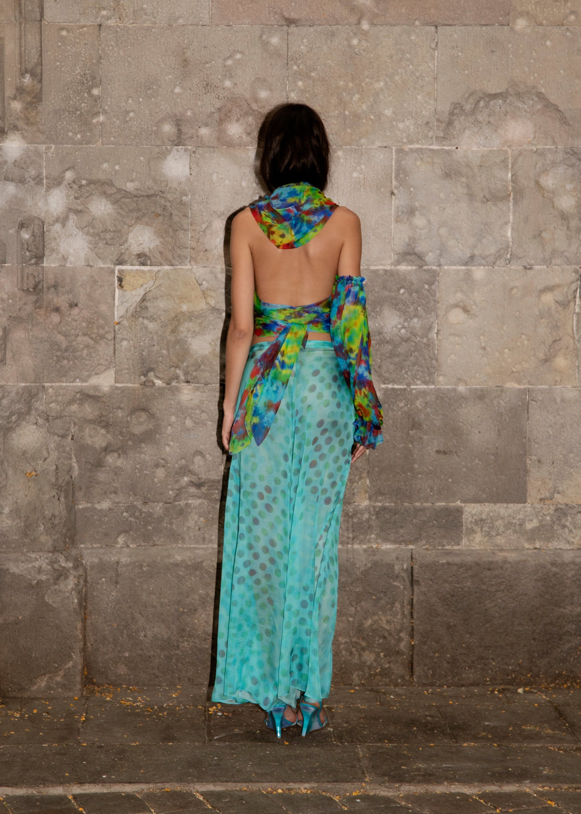Woman in Dramatica pants in acqua salata with colorful patterns against a stone wall.