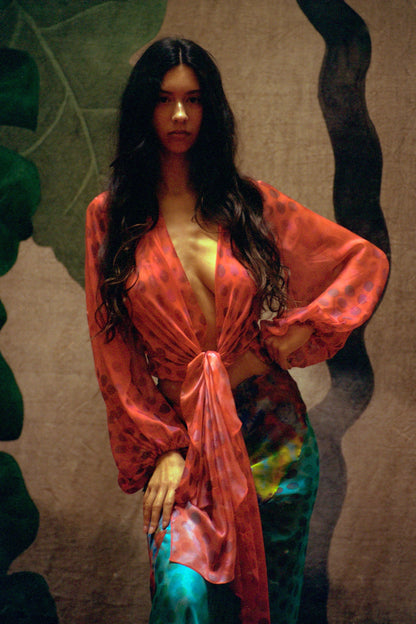 Woman wearing Electrica blouse in Cherry Sangria with a textured wall and leafy plant in the background
