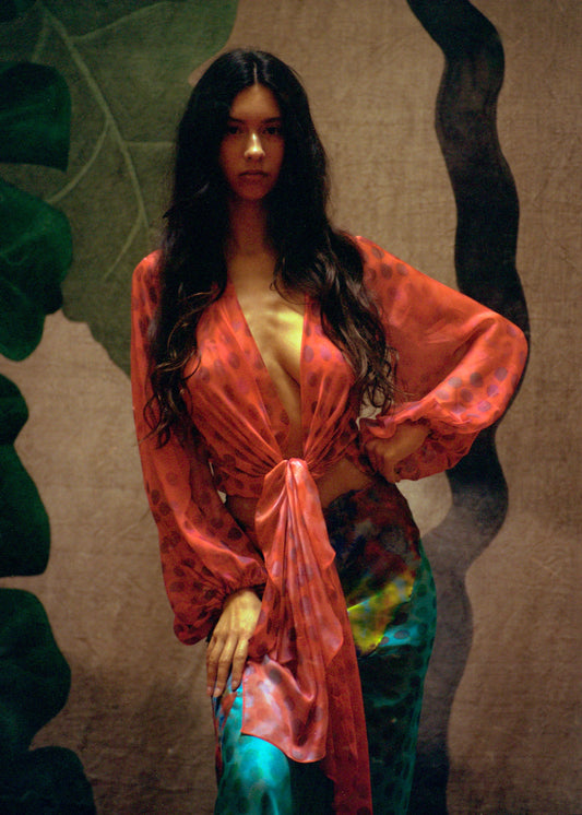 Woman wearing Electrica blouse in Cherry Sangria with a textured wall and leafy plant in the background