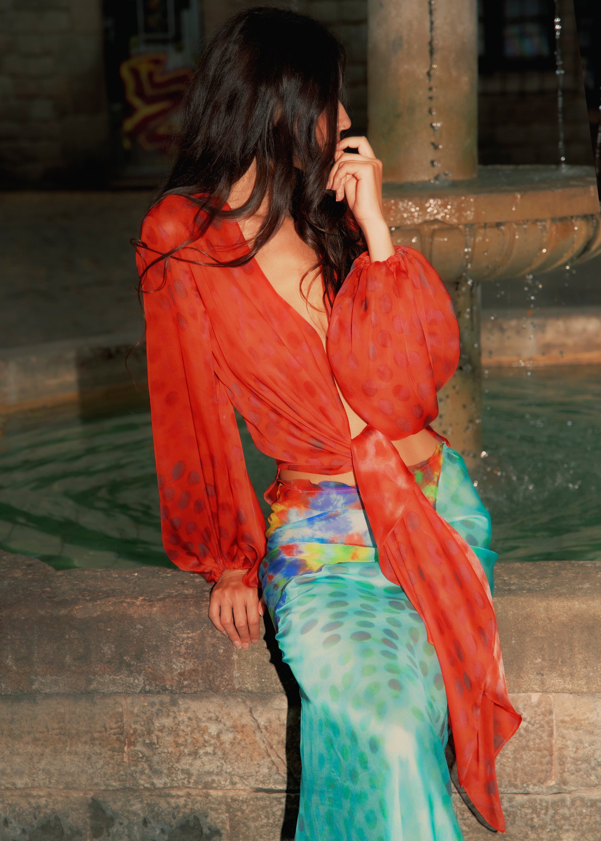 Woman in Electrica blouse in Cherry Sangria sitting by a fountain at night.