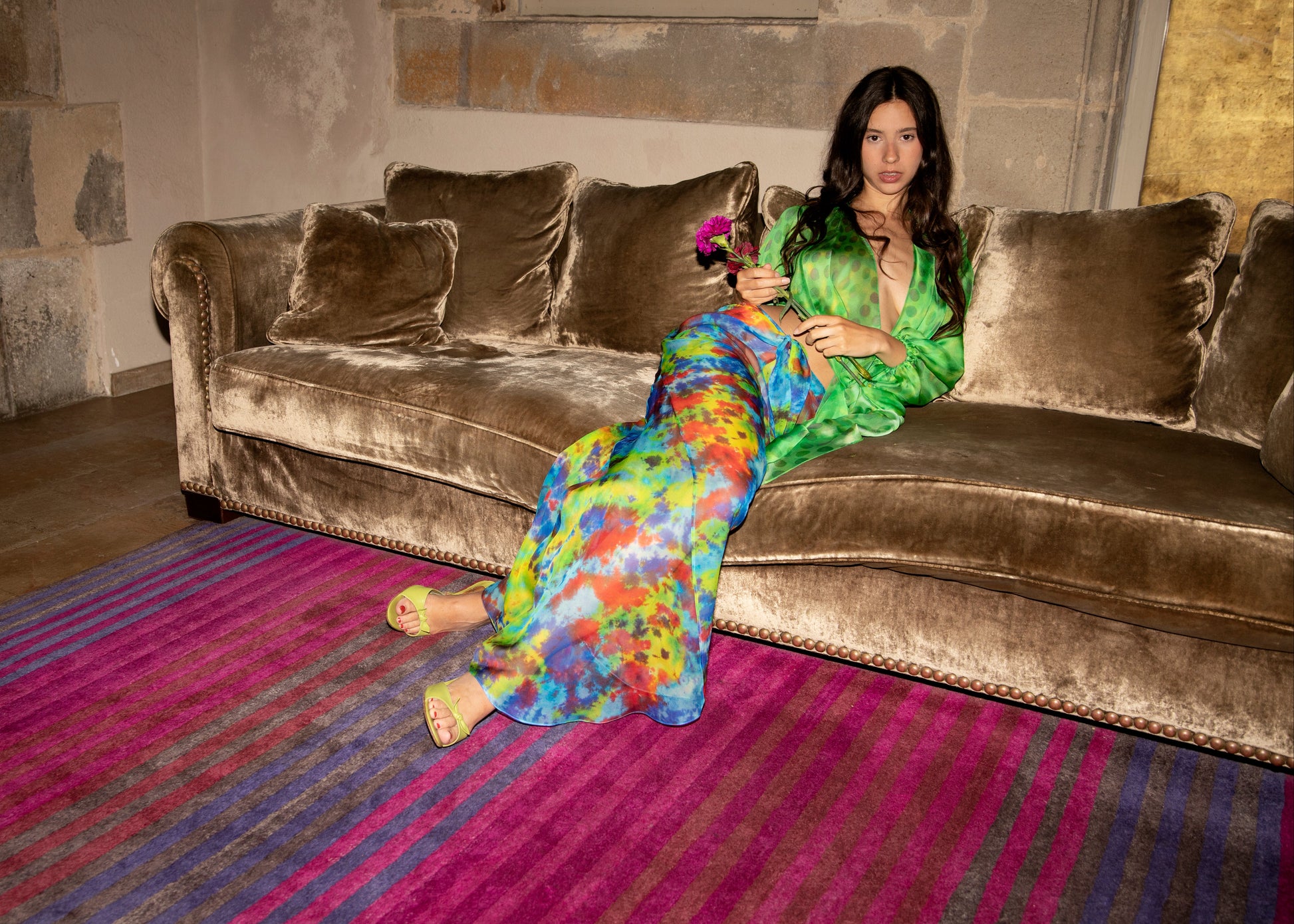 Woman in a colorful Dramatica pants in daydream sitting on a brown velvet sofa in a room with a striped rug.