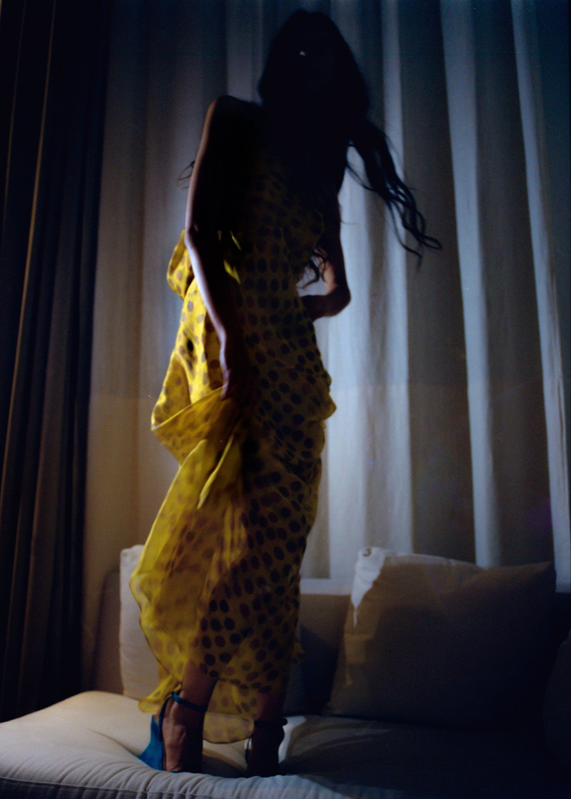 Woman in Romantica dress in Honeybaby  standing in a dimly lit room with curtains.